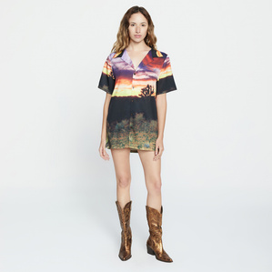 E-commerce catalogue image of female model, Jackie Reyes, wearing gold cowboy boots and an oversized short sleeve button shirt featuring print pattern of a dessert sunset standing in front of a white backdrop in-studio.