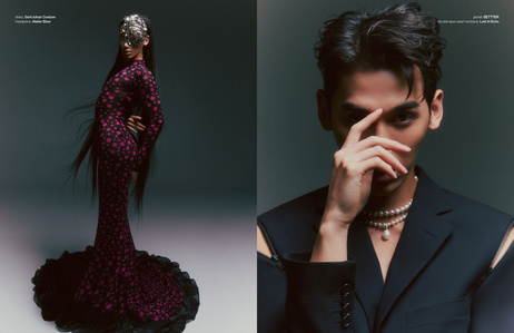 Plastique Tiara from RuPaul's Drag Race standing in front of a gray backdrop for a fashion editorial. She wears a silver mask, black dress with fuchsia polka dots, and a silver designer mask. Plastique out of drag as a man wearing a designer suit jacket.
