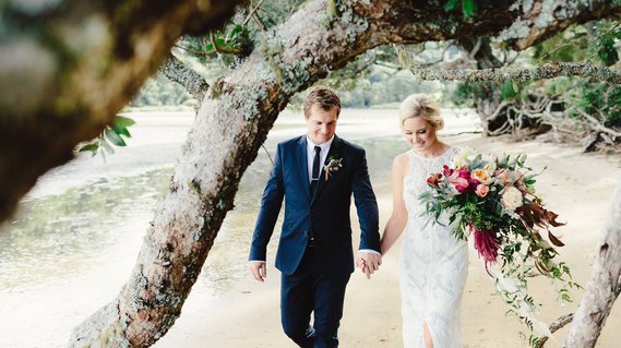 Blonde bride and groom on wedding day smiling and walking down beach at Opoutere, Coromandel, New Zealand