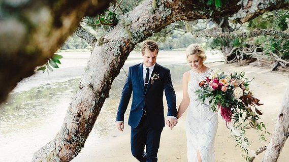 Blonde bride and groom on wedding day smiling and walking down beach at Opoutere, Coromandel, New Zealand