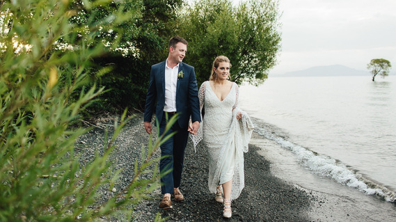 Smiling bride and groom walk along edge of Lake Taupo on their wedding day