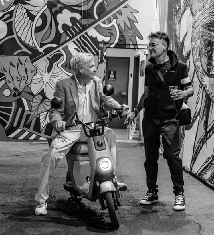 Peter Tunney on an electric scooter speaking with Adam Greenfader as they both share a cigar. 