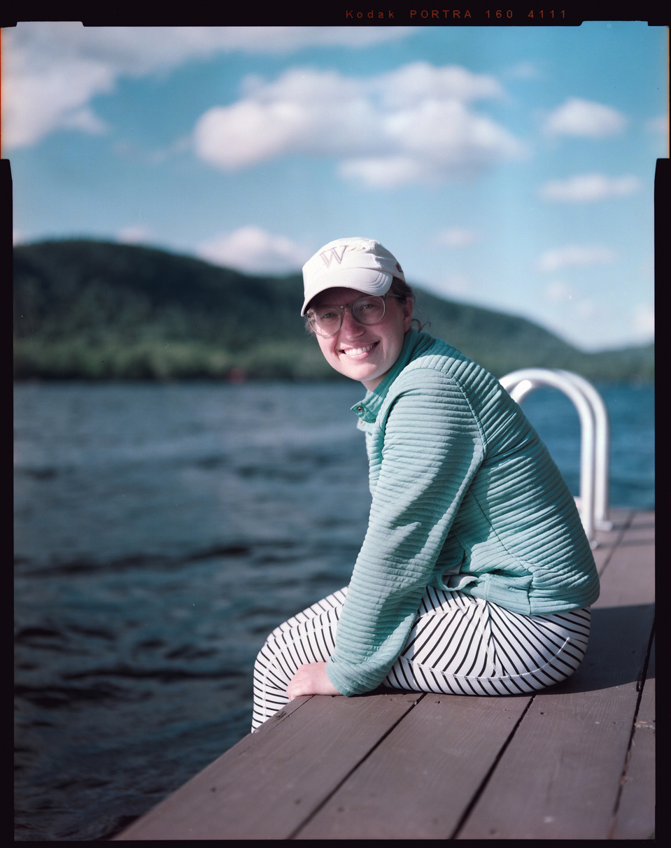 A young woman sits on a dock in a lake on a sunny day, looking at the camera. Mountains are behind her.