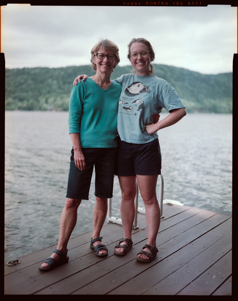 Two women stand together, the younger with her arm across the older's shoulder, looking at the camera. They are on a dock in a lake, with mountains behind them on a sunny day.