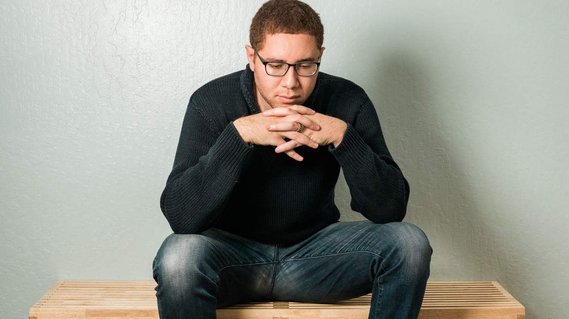 Ryan Jacobs, photographer. Self-portrait in indigo sweater and jeans, sitting on a bench, hands clasped.