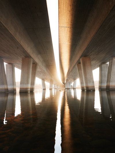 Bro af beton med vand under, hvor der er et flot lysindfald og en and som svømmer i midten. 