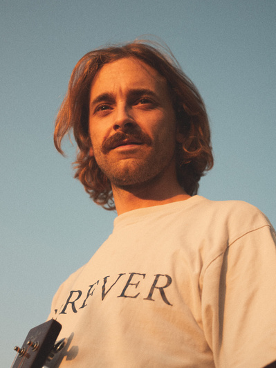 Musiker med langt hår står og nyder solnedgang med sin guitar og t-shirt hvor der står "forever".