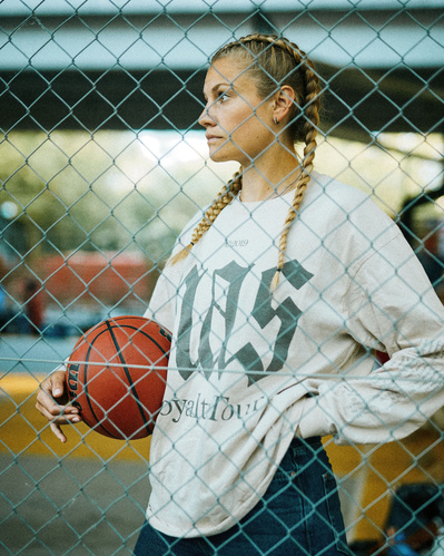 kvinde står bag et hegn med en basketbold under den ene arm og hånden i den anden lomme, hun har en trøje på med teksten "nas".