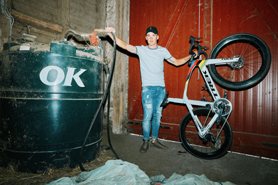 cykelrytter Mads Pedersen står ved en gammel benzintank og holder sin hvide racer cykel i den anden hånd