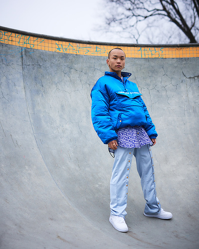 Skaldet asiatisk mand står på skatebane i blåt tøj og kigger ind i kameraet  