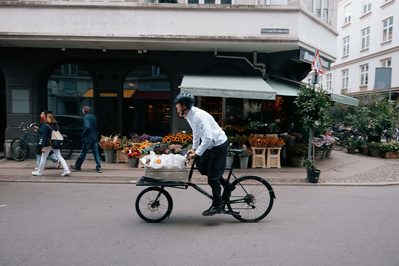 Mand cykler i byen på lang cykel med blomster på ladet og i hvid trøje.