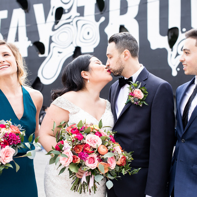 A couple dressed for their wedding share a kiss while their wedding party smiles at them