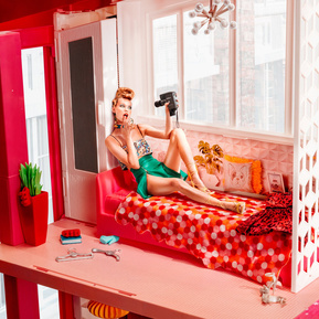 a barbie like woman is sitting in a barbie house with a vintage camera, shooting selfies