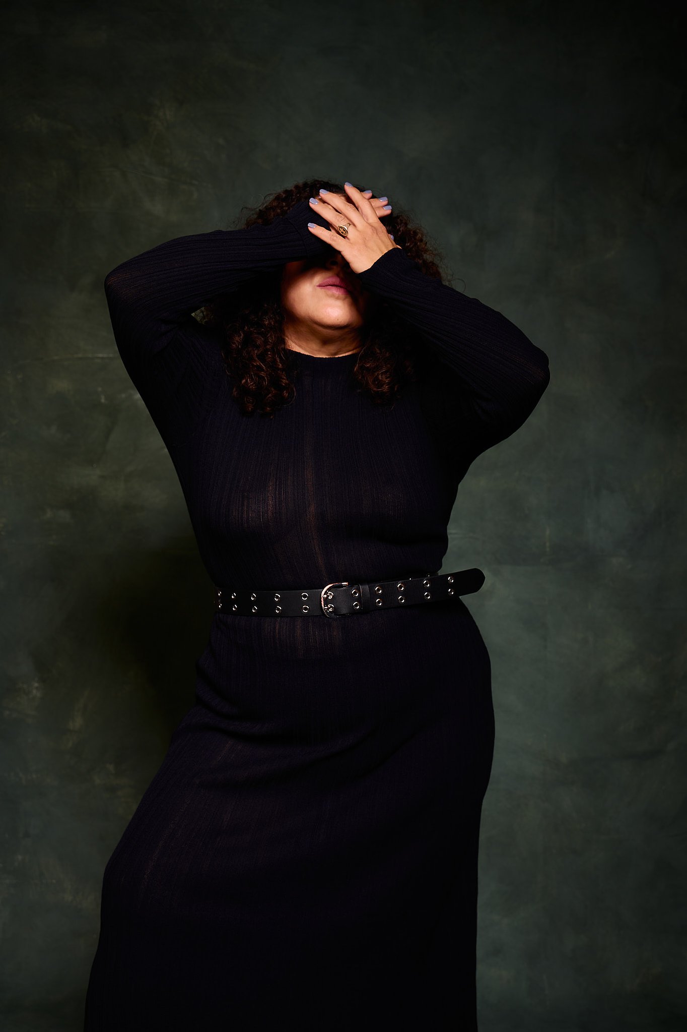 Retrato de mujer con pelo rizado y vestido transparencia negro en el estilo fotográfico de Boudoir. En pose cubriéndose el rostro. Fondo verde pintado a mano e iluminación de estudio.