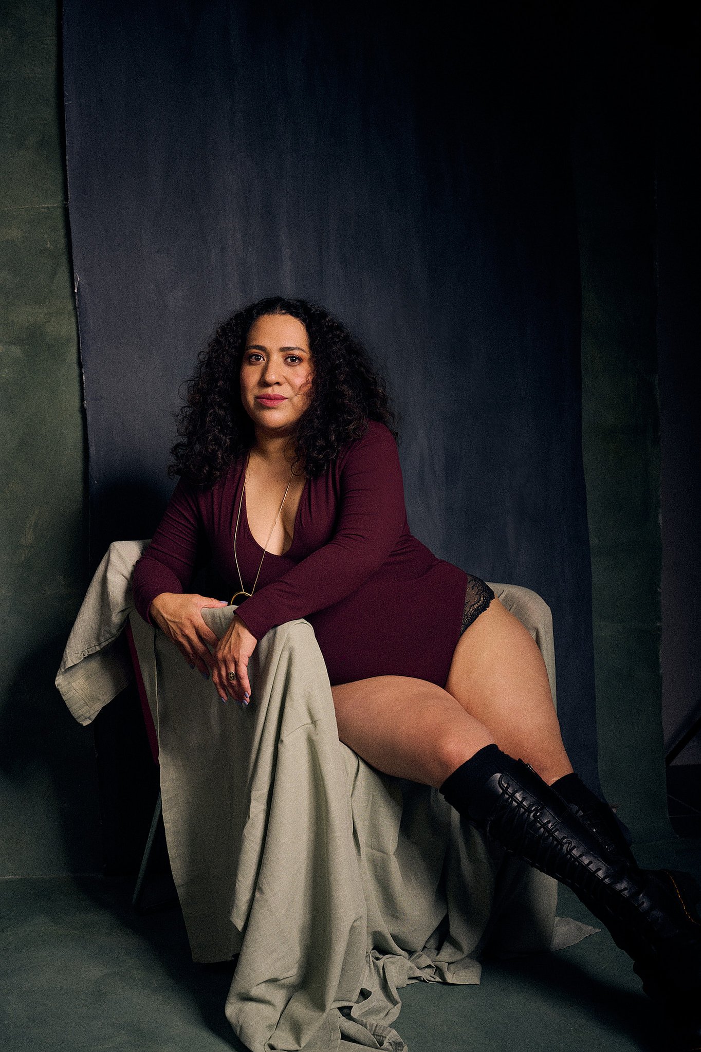 Retrato de mujer con pelo rizado y bodysuit rojo, con botas de piel a la rodilla en el estilo fotográfico de Boudoir. Fondo pintado a mano e iluminación de estudio.