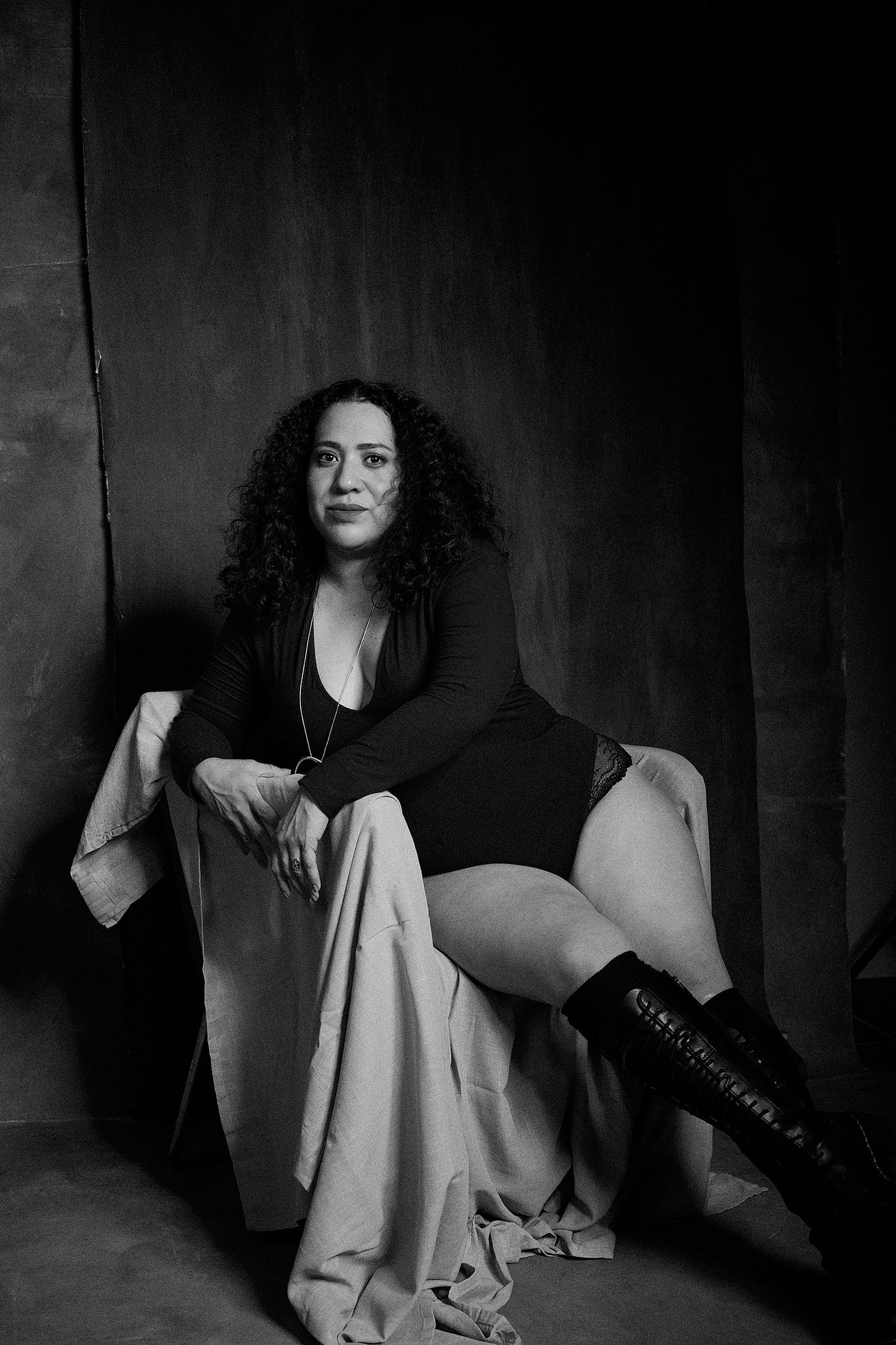 Retrato de mujer con pelo rizado y bodysuit negro, con botas de piel a la rodilla en el estilo fotográfico de Boudoir. Fondo pintado a mano e iluminación de estudio.