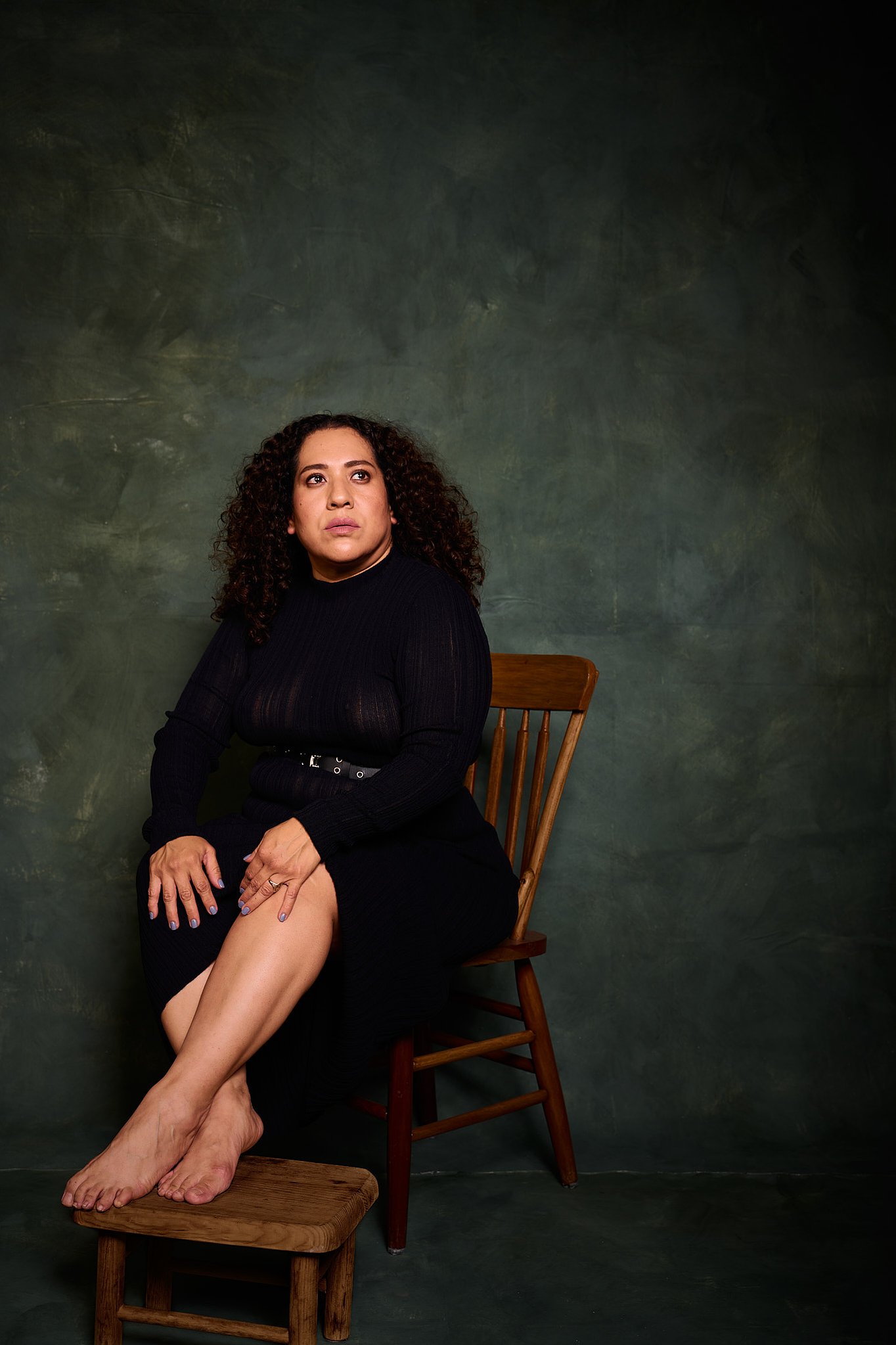 Retrato de mujer con pelo rizado y vestido transparencia negro en el estilo fotográfico de Boudoir, sentada en silla de madera y banco. Fondo verde pintado a mano e iluminación de estudio.