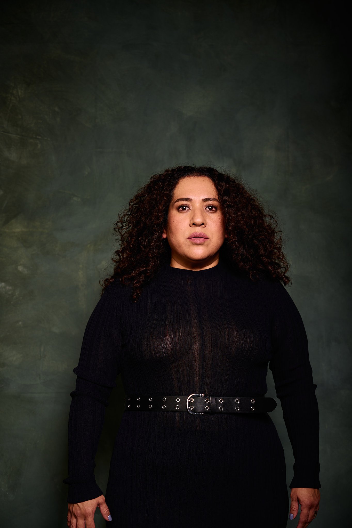 Retrato de mujer con pelo rizado y vestido transparencia negro en el estilo fotográfico de Boudoir. En pose de pie. Fondo verde pintado a mano e iluminación de estudio.