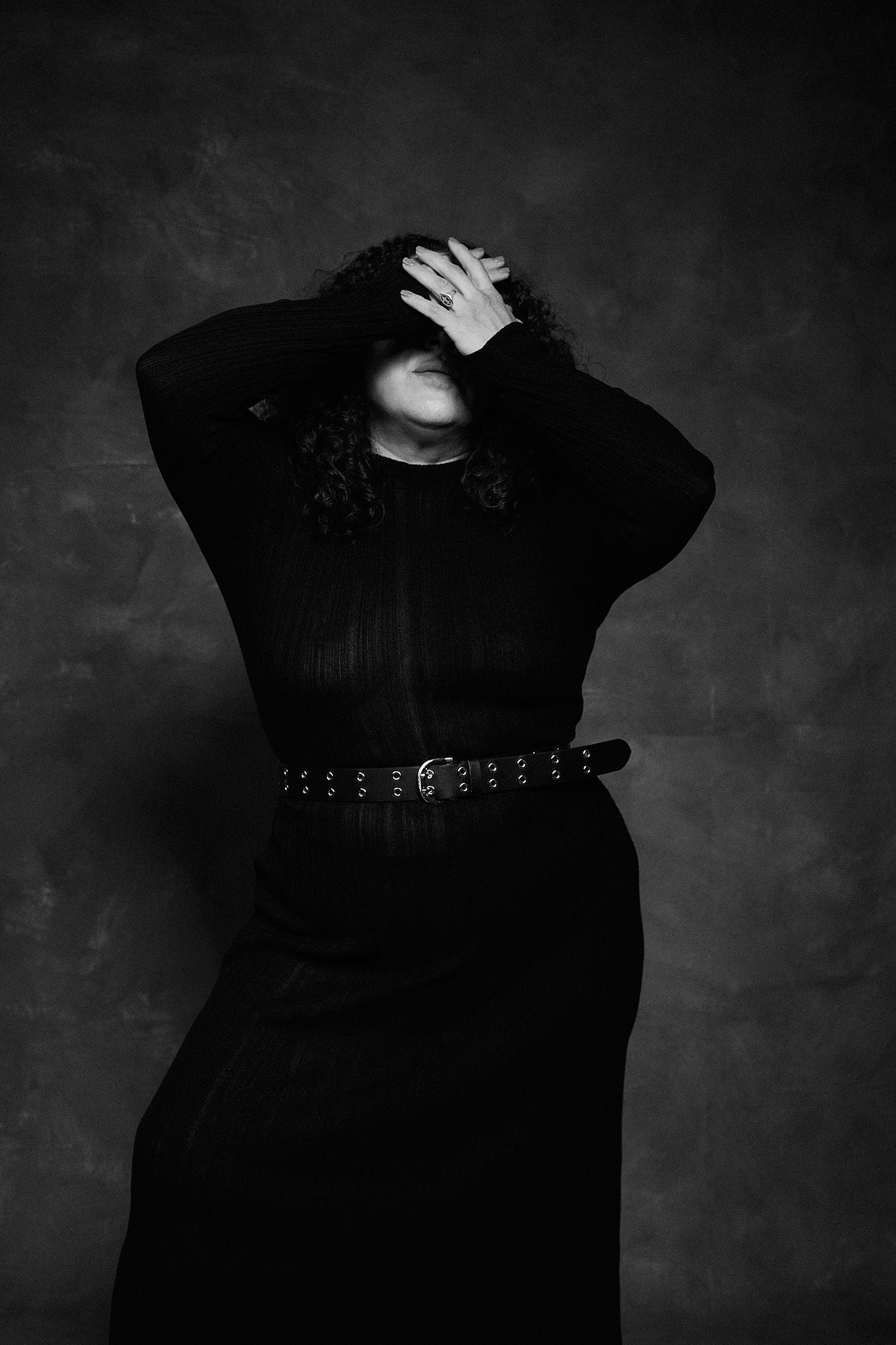 Retrato de mujer con pelo rizado y vestido de transparencia negro, en el estilo fotográfico de Boudoir.  Pose con manos cubriendo en rostro de forma dramática. Fondo pintado a mano e iluminación de estudio.