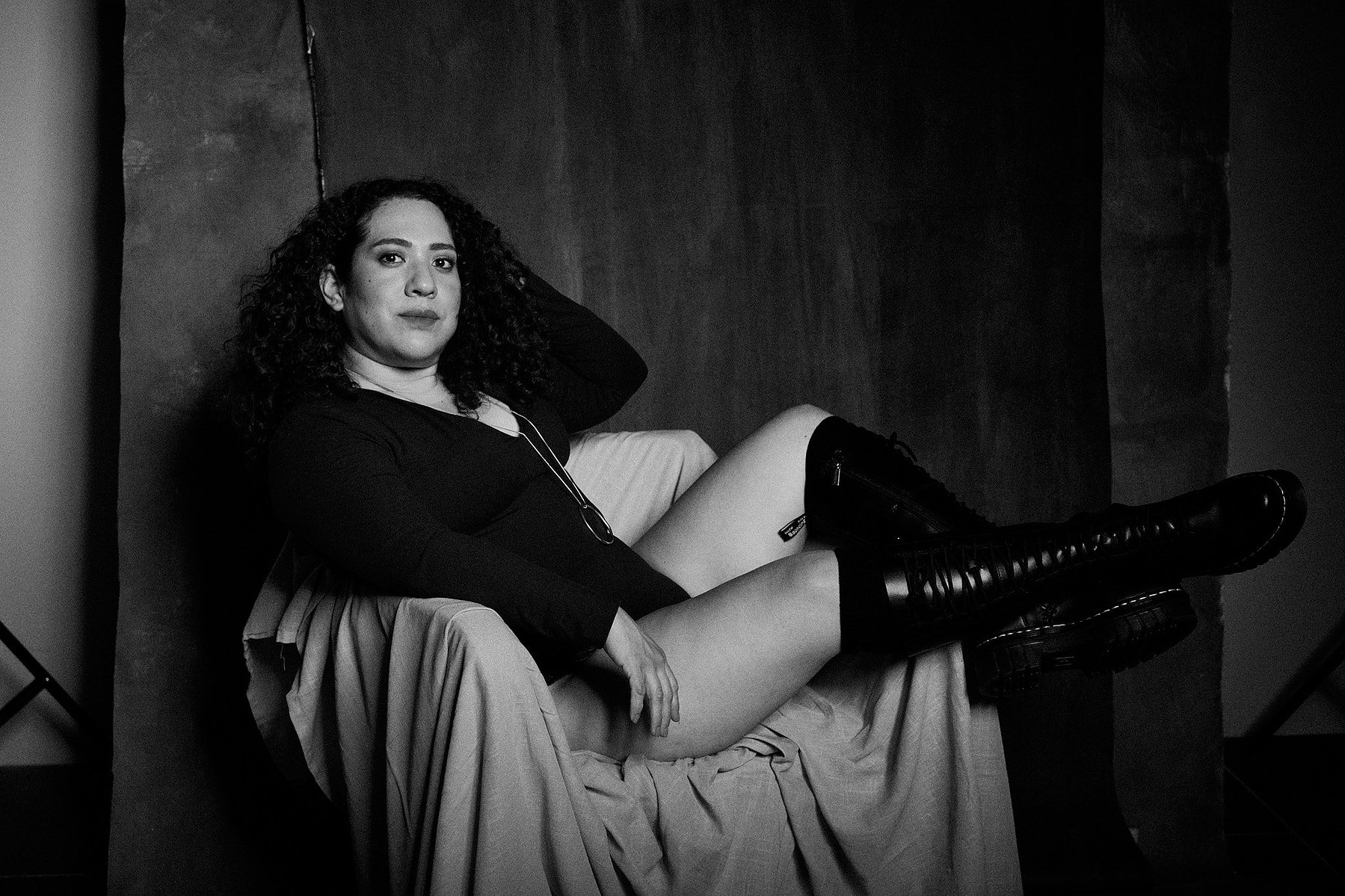 Retrato de mujer con pelo rizado y bodysuit negro, con botas de piel a la rodilla en el estilo fotográfico de Boudoir. Fondo pintado a mano e iluminación de estudio.