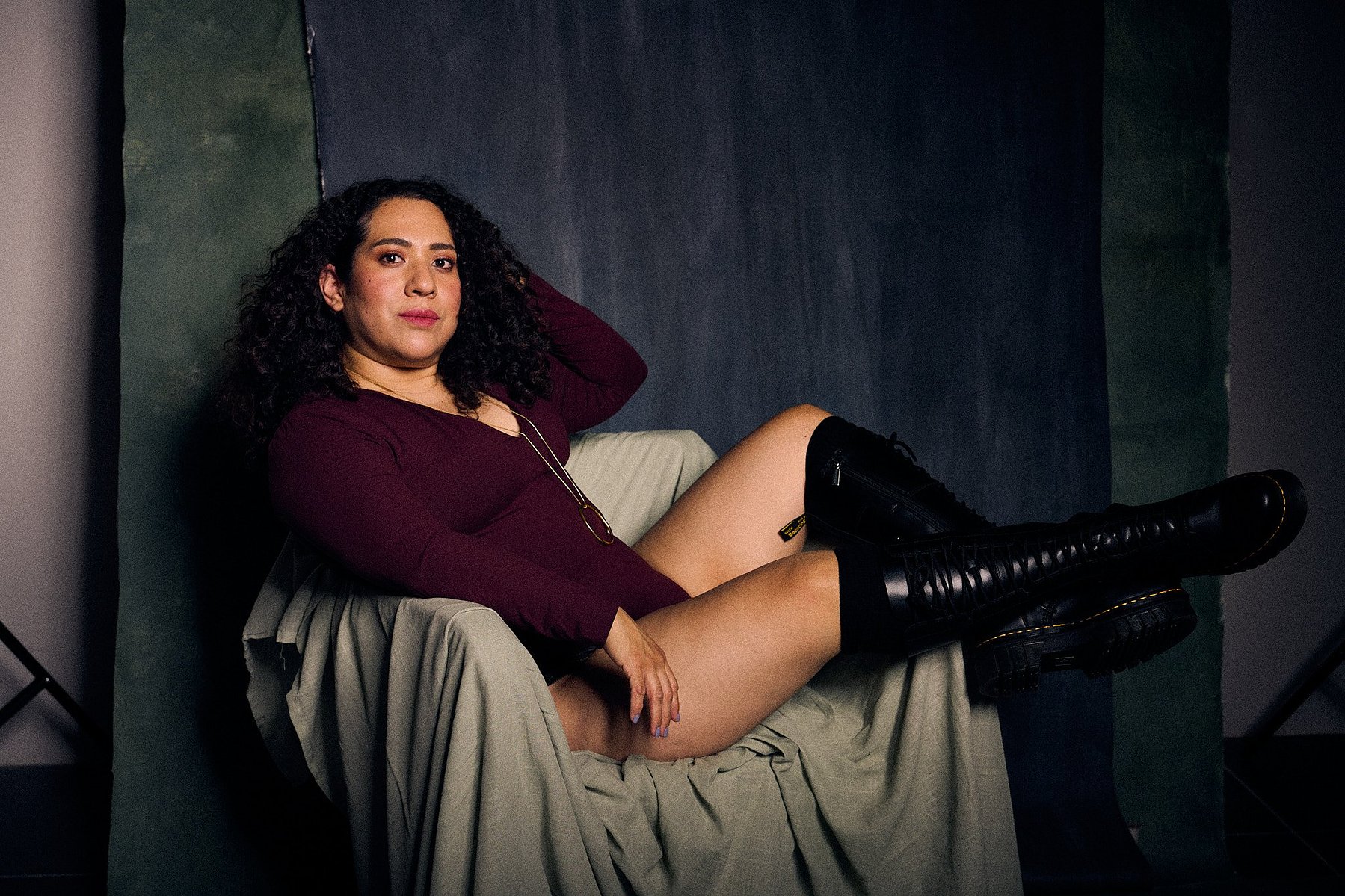 Retrato de mujer con pelo rizado y bodysuit rojo, con botas de piel a la rodilla en el estilo fotográfico de Boudoir. Fondo pintado a mano e iluminación de estudio.