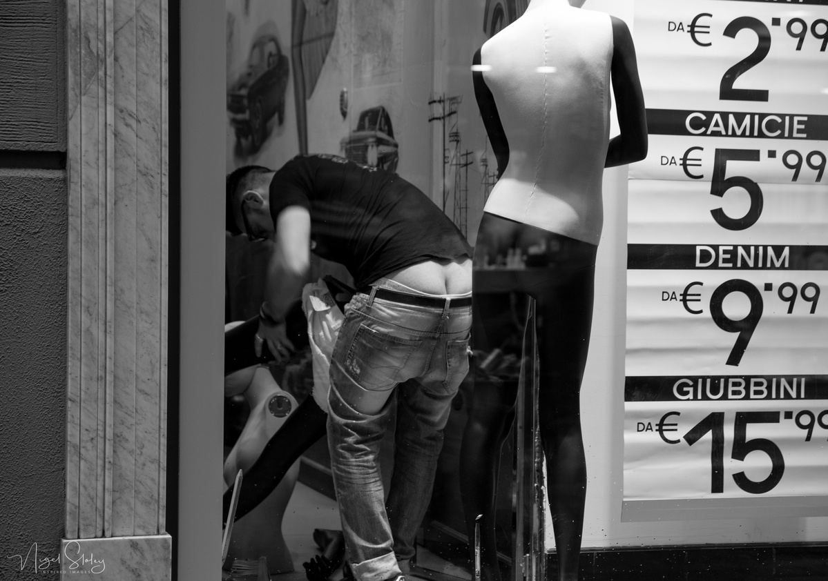 Black and white photograph shot in Naples, Italy. A window dresser with his but crack showing in the front window of a department store with a mannequin in a black dress. Nigel Sloley Photography
