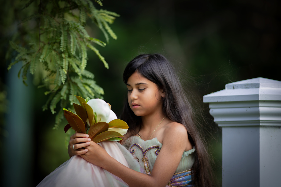 Portrait of a young girl wearing a pretty fairy gown sitting on a wall holding a large cream Magnolia flowers shot by Nigel Sloley Photography at Akatarawa Valley in New Zealand.