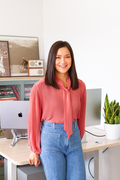Margaux Parker, a young graphic designer, stands in her brightly-lit office and leans back onto the edge of her desk. She rests her hands behind her on the desk and smiles widely at the camera.