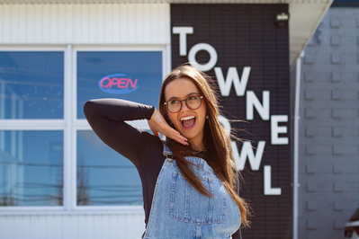 Young social media content creator Spencer Barbosa stands in front of Towne Bowl. The shot is framed from her waist up and she smiles with her mouth wide open while looking up and to the left. One arm is framing her face under her chin.