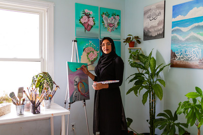 Hijabi young artist Hafsa Khizer stands in the middle of the frame holding an artist's palette in one hand and a paintbrush up to a painted canvas in the other. She looks solemnly at the camera. Paintings are on the wall behind her.