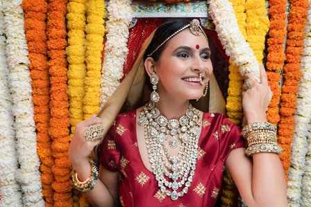 bride peeking out from a curtain of flowers wearing red designer dress and diamond jewellery shoot by best fashion photographer based in mumbai india