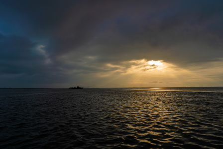 professional landscape photograph of a cloudy sky with the sun peaking through the clouds over an ocean with rough waves and a silhouette of a ship taken by leading landscape photographer ashish gurbani based in pune india