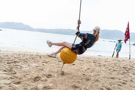 candid photograph of a female swinging on a yellow ball attached to a rope on a beach with hills and water in the background shot professionally by leading landscape photographer ashish gurbani based in pune india