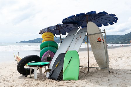 outdoor photograph of surf boards and tyres kept on a beach under two blue umbrellas with people hills water and blue sky in the background professionally shot by top landscape photographer ashish gurbani based in pune india