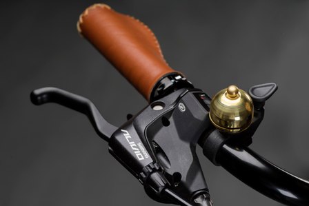 A product photograph of a handle bar of a black coloured cycle with a gold bell and a brown leather handle cover with a grey background shot in studio by best product photographer Ashish Gurbani based in Pune, India