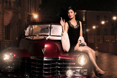 a professional photoshoot of a female model sitting on a red antique car  with buildings and lamp posts in the background shot by best luxury photographer ashish gurbani based in pune india