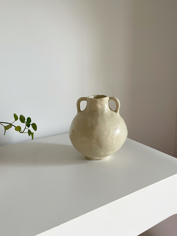a large round amphora vessel that is off-white sits on a white tabletop. to the left there is a plant that is slightly entering the photo.
