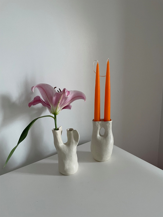 two white vessels sit on a white table. in the left vessel is a lilly flower and in the other vessel are two bright orange candle sticks