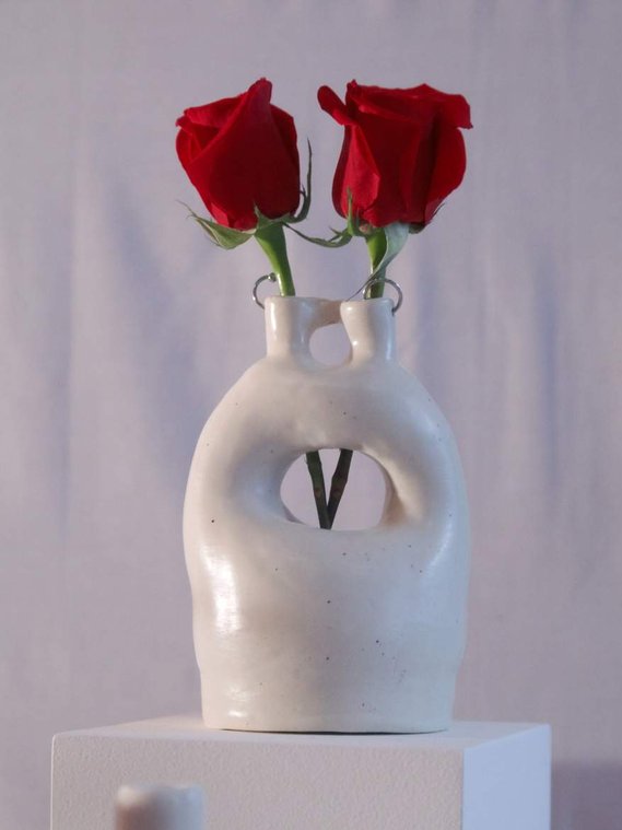an off-white vessel with a hole in the middle and by the top. the top is pierced with two silver hoops. there are two roses inside of this vessel.