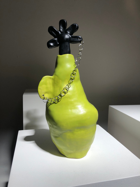 a lumpy bright lime green vessel with an ear-like handle. the handle is pierced and has a chain through it, which connects to the ceramic flower petal that is atop it.