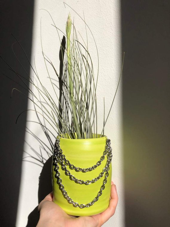 a  white hand holds a bright lime green vertical vase that has silver chains connected to it in three layers. an air plant is inside and stick out of the vase.
