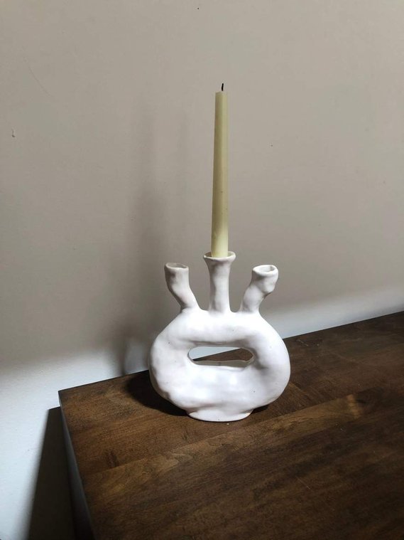 a three-candled candelabra sits on a brown wooden dresser with a white wall in the background. there is a yellow candle in the middle candle holder.