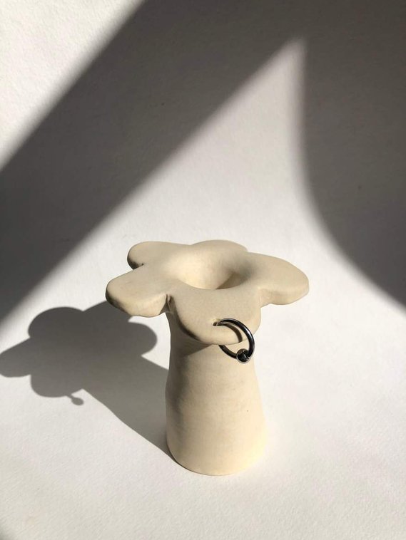 a vase that is off-white and has petal at the top of the stem like a flower. one petal is pierced with a silver hoop.