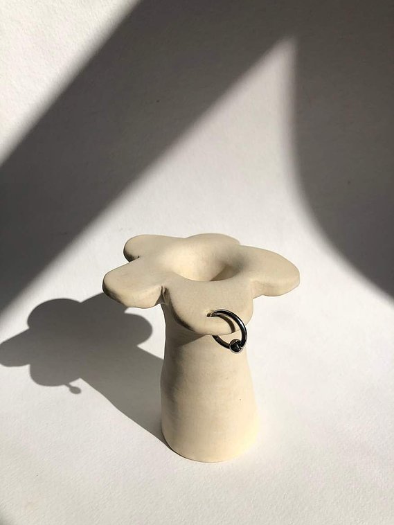 a vase that is off-white and has petal at the top of the stem like a flower. one petal is pierced with a silver hoop.