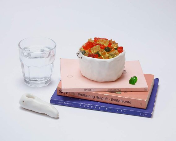 a white pipe sits next to a glass of water and a stack of three books. on top of the books is a white glazed pinch ceramic bowl with a silver hoop piercing it. the bowl is filled with gummy bears.