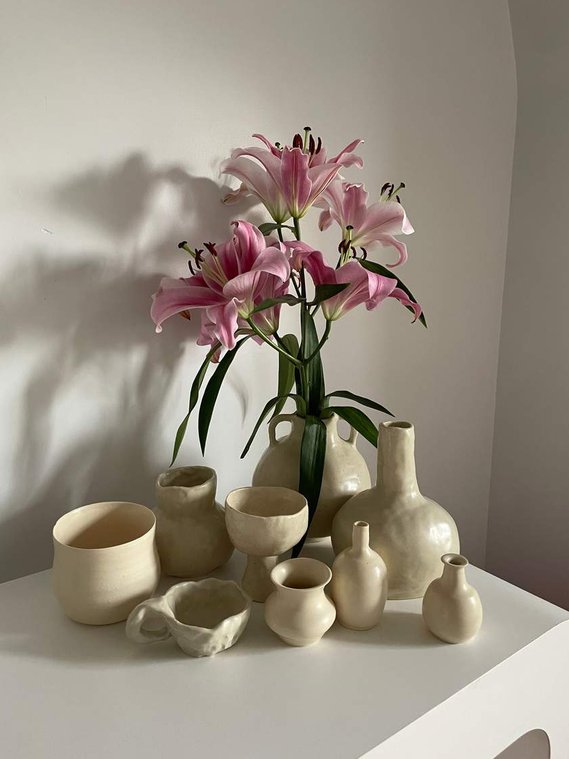 12 vessels in off-white sit on a white table. one vessel has pink lillies in the back.