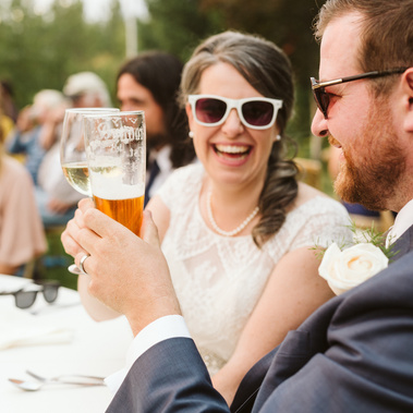 bride and groom wearing ray bans laughing and clinking beer mugs during their wedding reception in Glacier National Park, Montana.