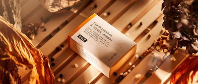 Frankincense and black pepper skincare soap product, placed on a light wooden backdrop along with raw peppercorns, dried plants and sea salt.
