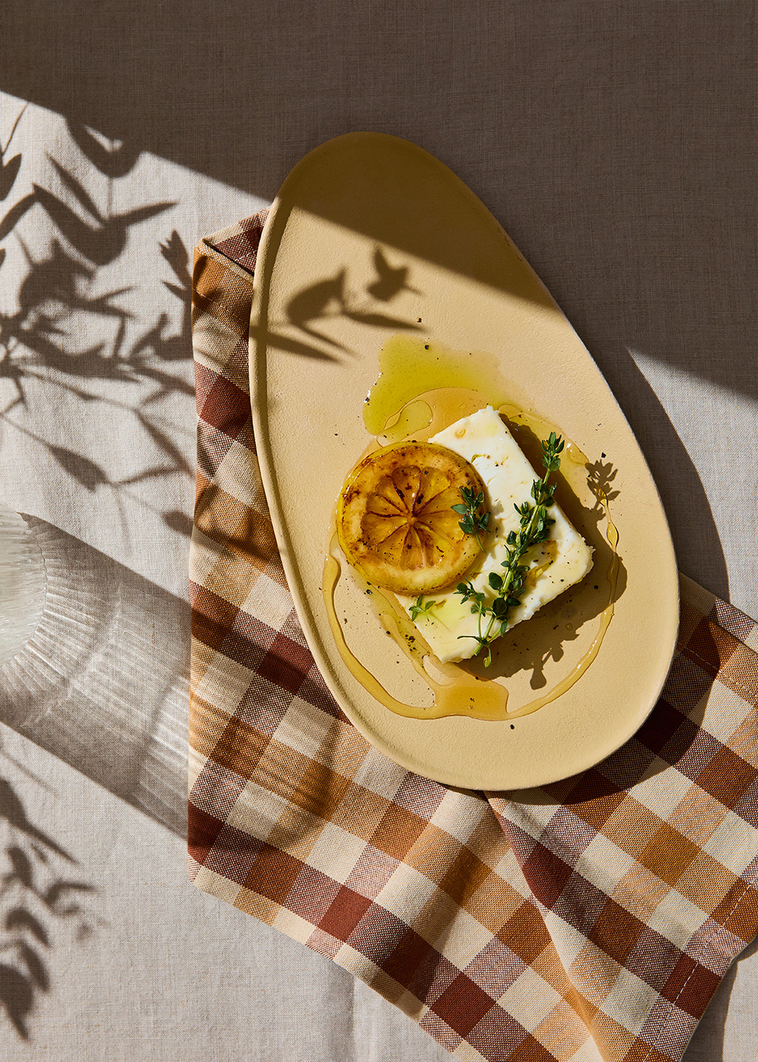 Hard sun drenches a muted yellow ceramic plate with crisp light and shadows. Featuring feta, griddled lemon slice thyme and olive oil. Set upon a gingham napkin. Photography by Lol Johnson. Food prep and styling by Iona Blackshaw. Props by Nuala Sharkey.