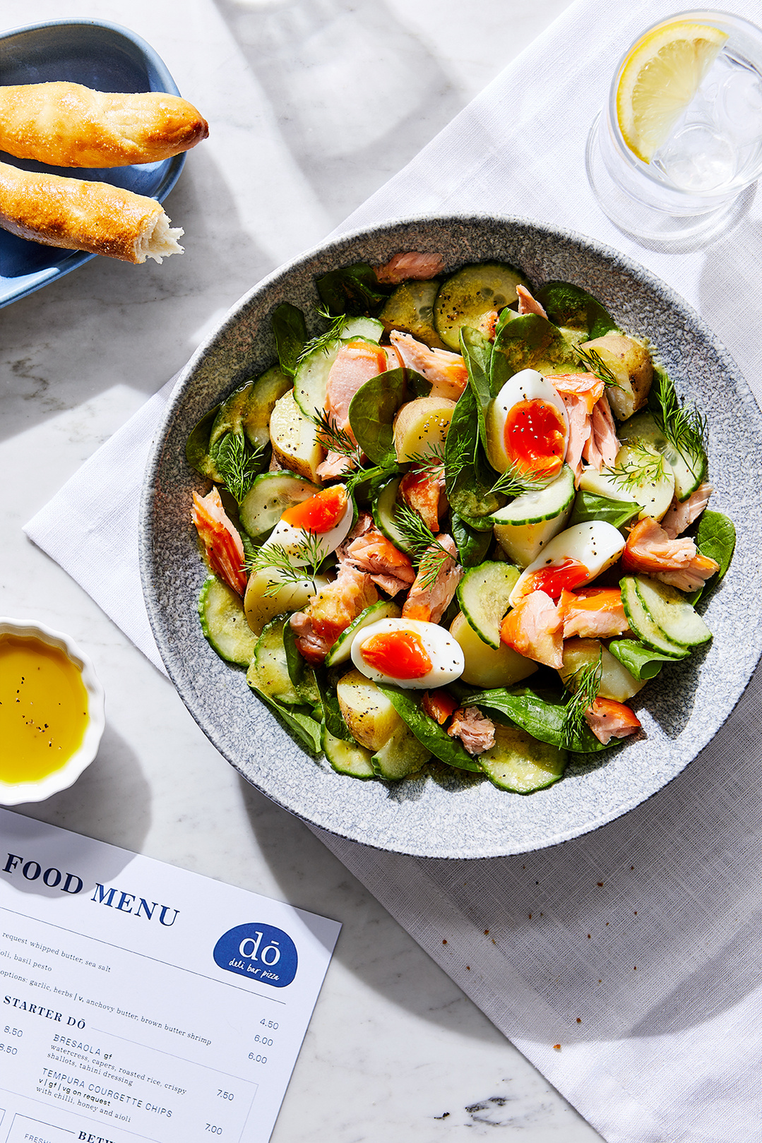 A sunny table with a hot smoked salmon and dill salad and dough sticks by Do Deli @ Hilton. Photography by south east London based food photographer Lol Johnson. Food prep and styling by Iona Blackshaw. Recipe development by consultant chef Jane Alty.